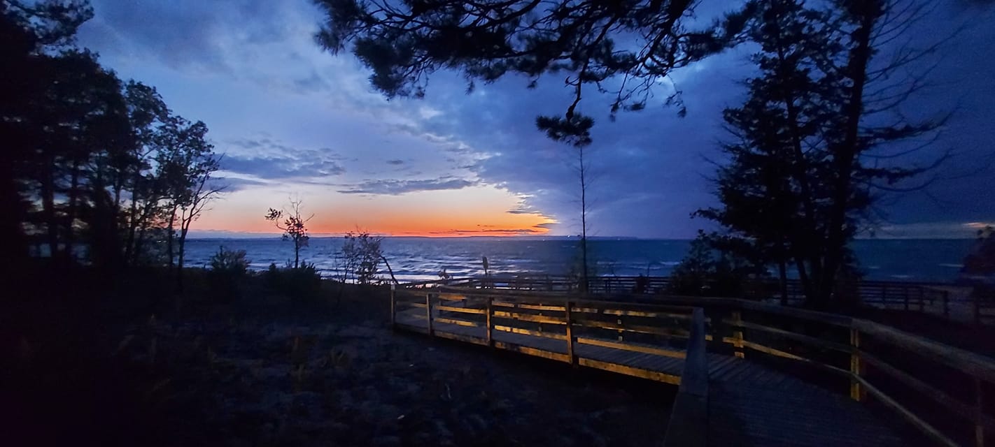 A boardwalk stretches towards the water, the sky is cloud filled except at the horizon, where its orange